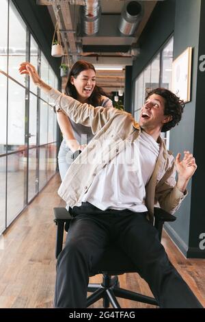 Junges Paar, das Spaß hat, als Frau den Mann auf dem Weg des modernen Großraumbüros in einem Stuhl schiebt Stockfoto