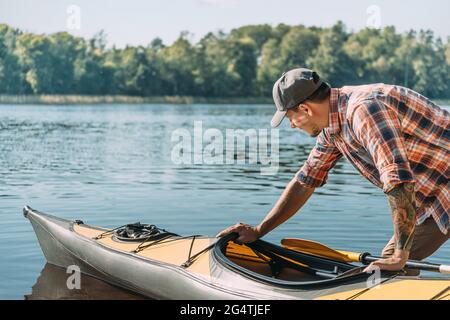 Junger Mann mit Mütze und Hemd schiebt ein Kajak im Wasser des Sees. Stockfoto