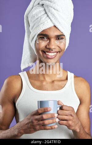 Junger Mann mit Augenflecken im Handtuch auf dem Kopf und einer Tasse Tee Porträt Stockfoto