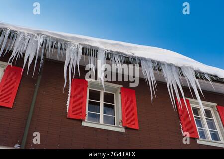 Große Eiszapfen hängen vom Dach und den Dachrinnen eines typischen Schweizer Hauses mit versperrten Fenstern in Engelberg, Zentralschweiz Stockfoto