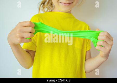 Hände mit grünem Schleim aus der Nähe. Ein Kind in einem gelben T-Shirt, das Schleim streckt. Trendy flüssiges Spielzeug klebt an den Händen. Kinder Hände spielen Schleim Spielzeug. Stockfoto