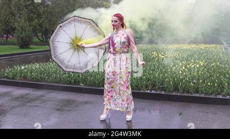 Entzückendes Mädchen in einem Kleid mit einem Blumendruck mit Regenbogen Zöpfen und Make-up. Tanzt mit einem Regenschirm, der sich in gelbem Rauch vor der Kulisse von FL Stockfoto