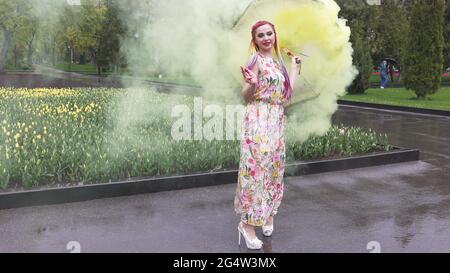 Entzückendes Mädchen in einem Kleid mit einem Blumendruck mit Regenbogen Zöpfen und Make-up. Tanzt mit einem Regenschirm, der sich in gelbem Rauch vor der Kulisse von FL Stockfoto