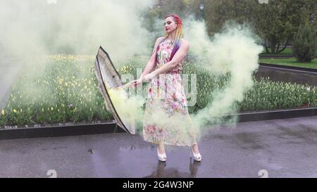 Entzückendes Mädchen in einem Kleid mit einem Blumendruck mit Regenbogen Zöpfen und Make-up. Tanzt mit einem Regenschirm, der sich in gelbem Rauch vor der Kulisse von FL Stockfoto