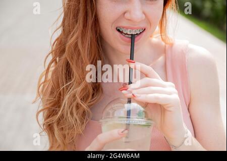 Junge schöne rothaarige Frau lächelt mit Zahnspangen Getränke kühlen Limonade im Sommer im Freien. Cropped Portrait Mädchen mit Sommersprossen Stockfoto