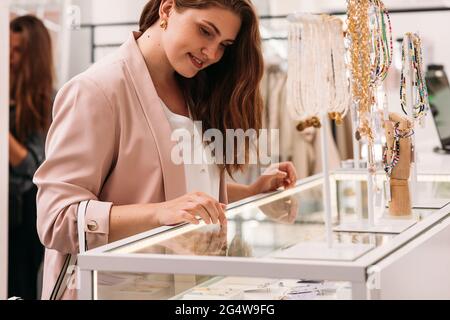 Junge elegante Frau, die auf Accessoires im Vitrine schaut. Lächelnde Käuferin, die in einem Bekleidungsgeschäft an einer Theke steht. Stockfoto