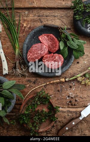 3 rohe Filet-Mignon-Steaks in grauem Steinteller auf rustikalem Holztisch mit Steakmessern und frischen grünen Kräutern Draufsicht Stockfoto