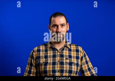 Porträt eines hübschen bärtigen Mannes in einem gelben karierten Hemd isoliert auf blauem Studiohintergrund. Posing-Konzept Stockfoto