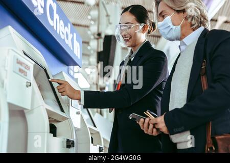 Eine weibliche Flugbegleiterin zeigt dem männlichen Passagier am Flughafenterminal das Self-Check-in-Verfahren an einem Automaten. Bodenpersonal trägt Gesicht s Stockfoto