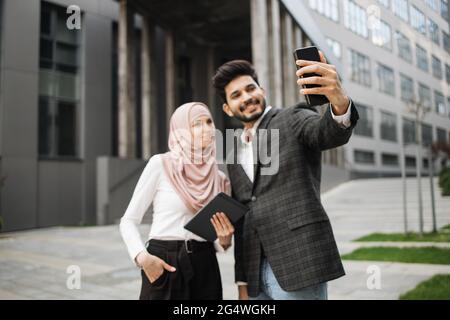 Attraktive Frau in Hijab hält digitale Tablette in der Nähe ihrer muslimischen Kollegen, die mit modernen Smartphone für Selfie zu nehmen stehen. Konzept von Business, Menschen und Gadgets. Stockfoto