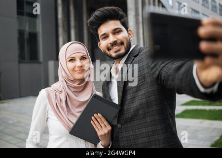 Attraktive Frau in Hijab hält digitale Tablette in der Nähe ihrer muslimischen Kollegen, die mit modernen Smartphone für Selfie zu nehmen stehen. Konzept von Business, Menschen und Gadgets. Stockfoto