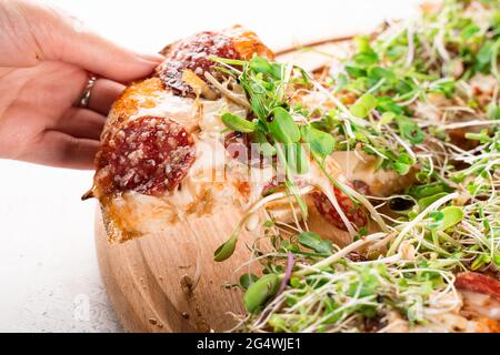 Stück Pinsa romana in der Hand mit streckenden Mozzarella mit Salami, Käse, Pilzen, verziert mit Microgreens auf Holzbrett. Stockfoto
