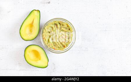 Zwei Avocado-Hälften und eine Glasschale mit vorbereiteter Guacamole auf einem Schreibtisch mit weißem Stein, Blick von oben für Text rechts Stockfoto