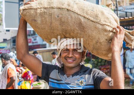 Lächelnder indischer Portier, der einen schweren Sack von Produkten auf dem Kopf trägt, Udupi, Karnataka, Indien Stockfoto