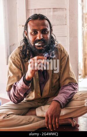 Nahaufnahme Porträt von Rikscha-Fahrer in Khaki Uniform bläst Rauch von Zigarette sitzend mit gekreuzten Beinen auf dem Boden des Cafés, Mysore, Karnataka, Indien Stockfoto