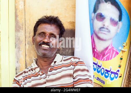 Nebeneinander - Porträt eines lächelnden indischen Mannes mit Schnurrbart, der neben dem Poster eines jungen indischen Mannes mit Schnurrbart sitzt, Mysore, Karnataka, Indien Stockfoto