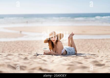 Junge Frau mit Buch, das wegschaut, während sie im Urlaub am Strand auf Sand liegt Stockfoto