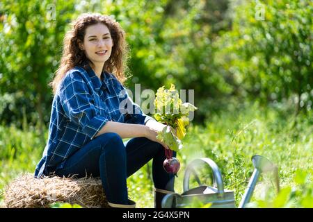 Lächelnde junge Frau, die auf einem Heustapel sitzt, während sie rote Beete hält Stockfoto