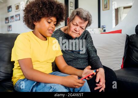 Ältere Frau, die den Enkel mit einem Smartphone ansieht, während sie auf dem Sofa im Wohnzimmer sitzt Stockfoto