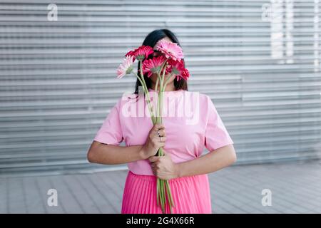Junge Frau bedeckt das Gesicht mit Gerbera-Gänseblümchen vor Wellblech Stockfoto
