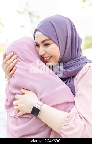 Lächelnde Frau, die in der Stadt einen Hijab trägt und sich einer Freundin umarmt Stockfoto