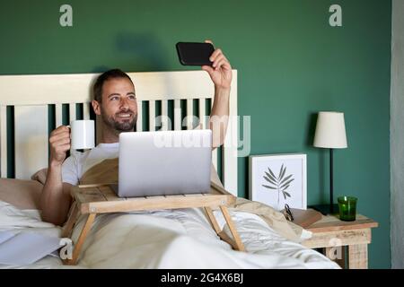 Lächelnder Mann mit Kaffeetasse, der Selfie über ein Smartphone im Schlafzimmer gemacht hat Stockfoto