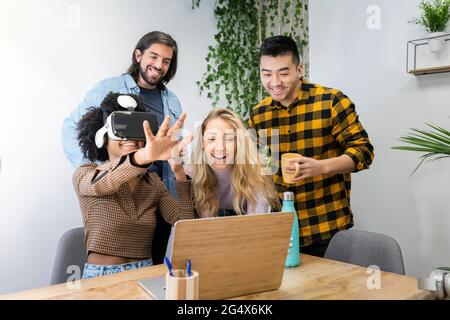 Geschäftsfrau im Virtual-Reality-Simulator von männlichen und weiblichen Kollegen, die im Büro einen Videoanruf über einen Laptop tätigen Stockfoto