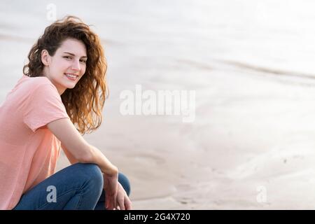 Lächelnde junge Frau, die am Strand auf Sand hockend Stockfoto