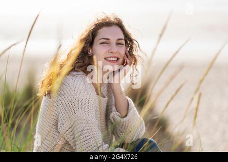 Lächelnde junge Frau mit der Hand am Kinn, die in den Dünen am Strand sitzt Stockfoto