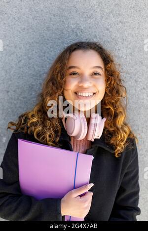 Lächelnde junge Frau mit Kopfhörern, die die Datei an die Wand halten Stockfoto