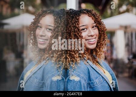 Lockige Frau in Denim-Jacke, die an einer Glaswand angelehnt ist Stockfoto