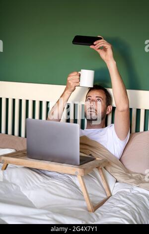 Ein Mann mittleren Erwachsenen, der im Schlafzimmer Selfie mit einer Kaffeetasse über ein Smartphone gemacht hat Stockfoto