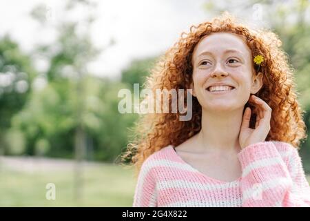 Lächelnde Frau mit lockigen Haaren, die Blume trägt, während sie wegschaut Stockfoto