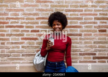 Lächelnde junge Frau mit wiederverwendbarem Becher, der vor einer Ziegelwand steht Stockfoto