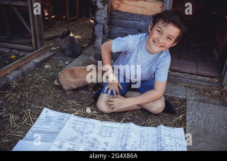 Lächelnder Junge mit Blaupause, der neben dem Kaninchen im Hinterhof sitzt Stockfoto