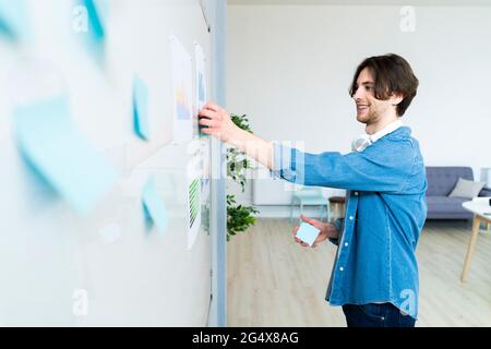 Lächelnder männlicher Unternehmer klebt Haftnotiz auf Whiteboard im Büro Stockfoto