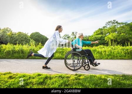 Sorglose weibliche Hausmeisterin, die im Rollstuhl im öffentlichen Park mit älteren Frauen Spaß hat Stockfoto