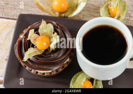 Nahaufnahme von Minipie mit Mascarpone-Creme und Schokoladenganache, dekoriert mit Physalis, neben einer Tasse Kaffee. Stockfoto