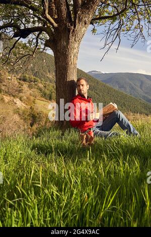 Ein hübscher Mann schaut weg, während er unter dem Baum auf dem Gras sitzt Stockfoto