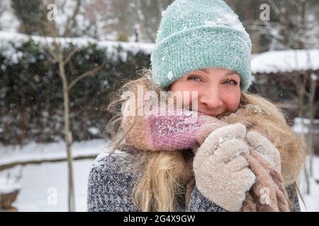 Blonde Frau bedeckt den Mund mit Schal, während sie während des Schnees im Hinterhof steht Stockfoto