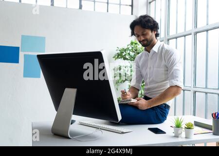 Männlicher Unternehmer, der auf einem Notizblock schreibt, während er auf dem Schreibtisch sitzt Stockfoto