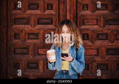 Lockige Frau, die den wiederverwendbaren Becher mit einem Smartphone an einer Holztür hält Stockfoto