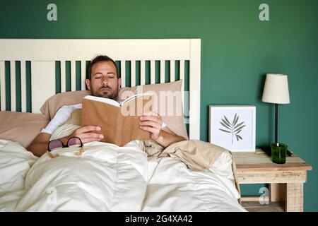 Mann mit mittlerem Erwachsenenalter liest Buch, während er sich im Schlafzimmer ausruhte Stockfoto