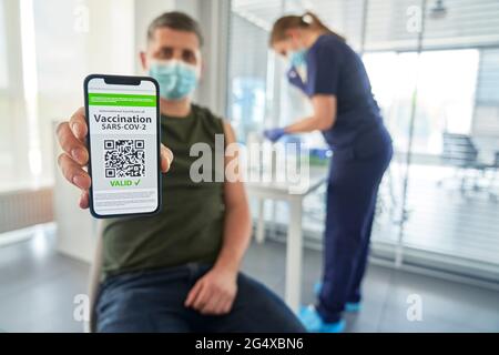 Mann, der den QR-Code für die COVID-19-Impfung auf dem Mobiltelefon zeigt, während der Arzt im Hintergrund in der Klinik war Stockfoto