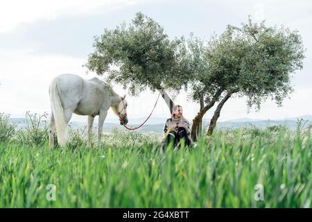 Junge Frau schaut weg, während sie unter dem Baum an einem Pferd sitzt Stockfoto