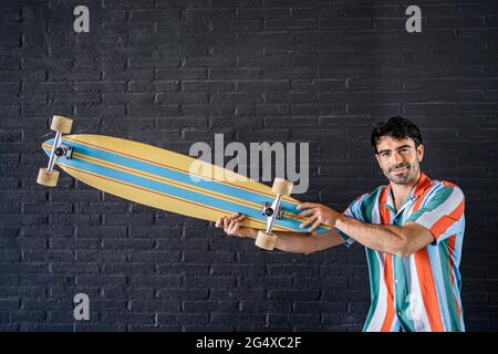 Junger, hübscher Mann, der Skateboard vor einer schwarzen Ziegelwand hält Stockfoto