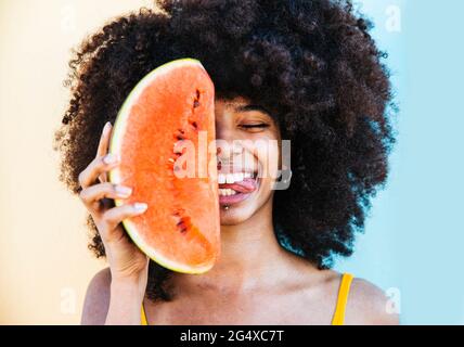 Verspielte Frau, die die Zunge herausstreckt, während sie die Wassermelonenscheibe vor dem Gesicht hält Stockfoto