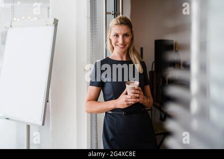 Lächelnde Unternehmerin mit wiederverwendbarer Kaffeetasse, die vor der Tür im Büro steht Stockfoto