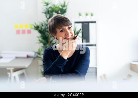 Lächelnde professionelle Frau, die im Büro über einen Lautsprecher auf dem Smartphone spricht Stockfoto