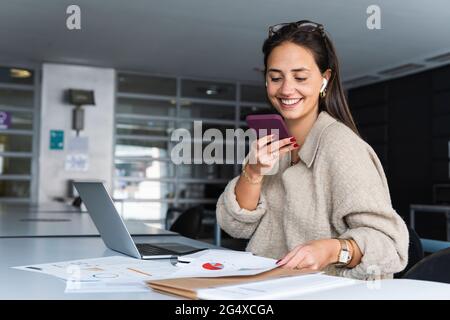 Lächelnde Geschäftsfrau, die im Büro Voicemail über das Mobiltelefon sendet Stockfoto